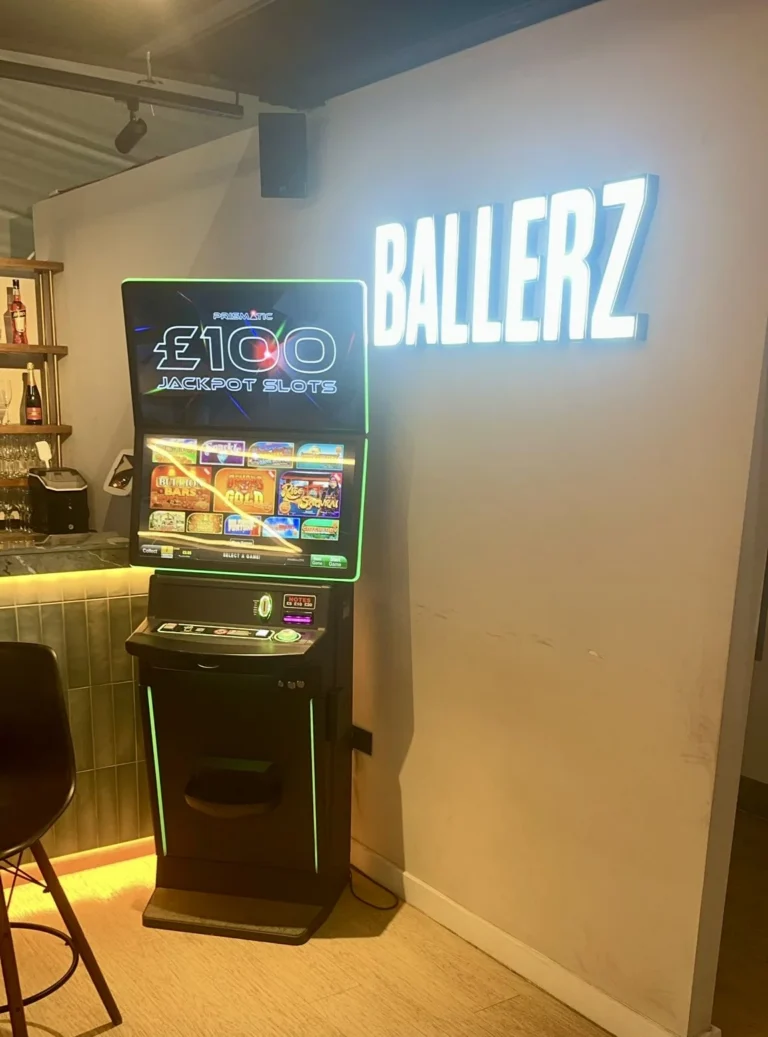 Slot machine in a bar showing £100 Jackpot Slots, with neon green trim and a glowing BALLERZ sign on the wall behind it.