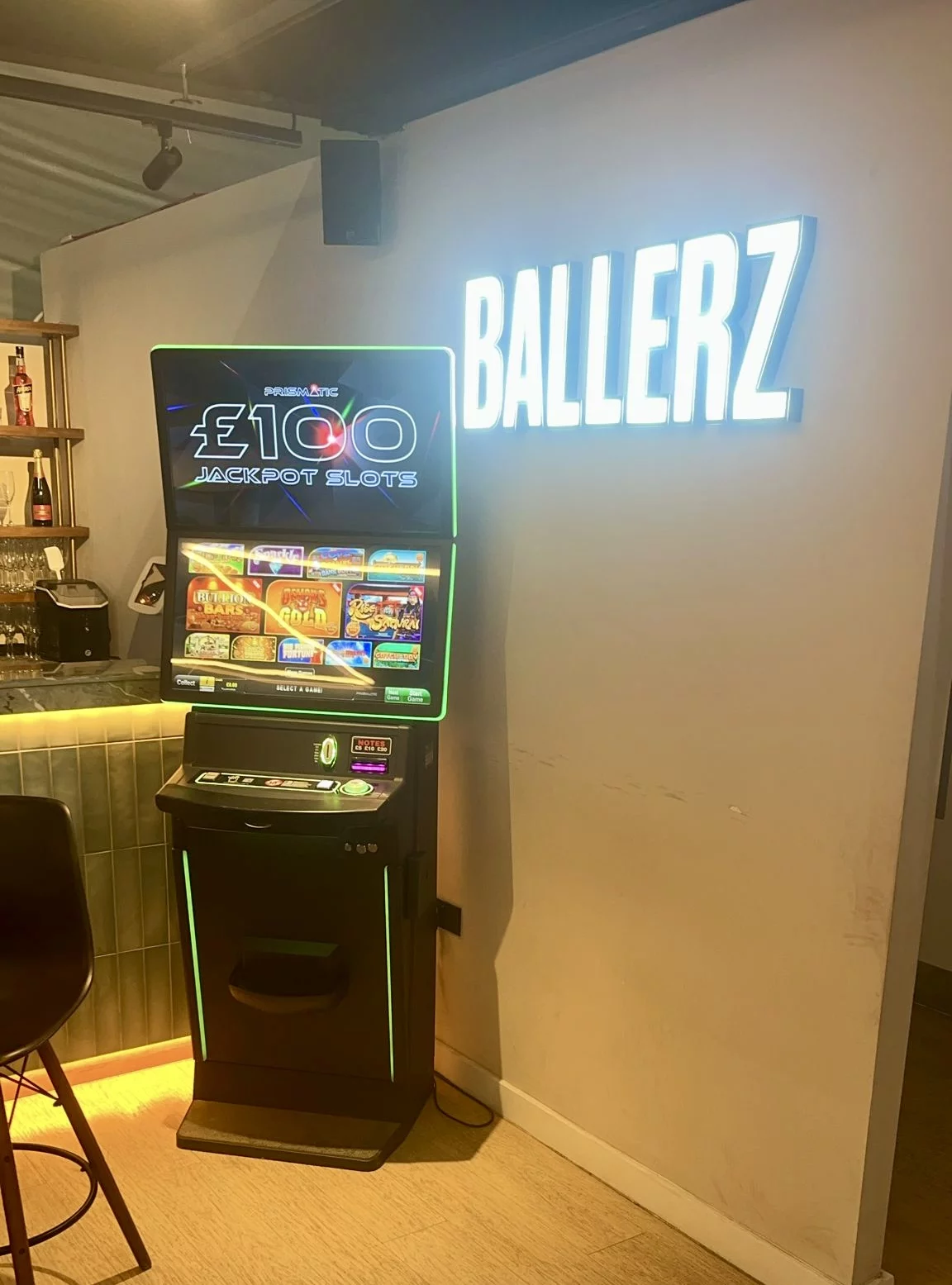 Slot machine in a bar showing £100 Jackpot Slots, with neon green trim and a glowing BALLERZ sign on the wall behind it.
